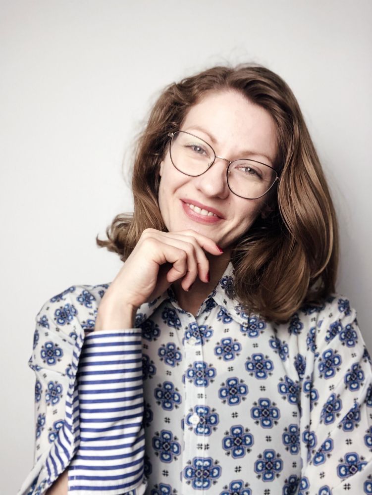 Headshot of Patrycja Wardal, a psychologist with wavy hair and glasses, smiling with her hand under her chin against a light background.